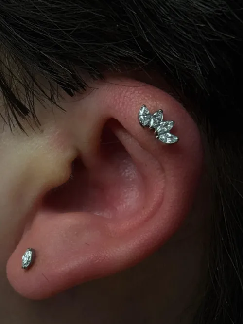 Prim-plan al unei urechi cu piercing helix decorat cu o bijuterie argintie cu cristale în formă de frunze și un piercing discret în lob.